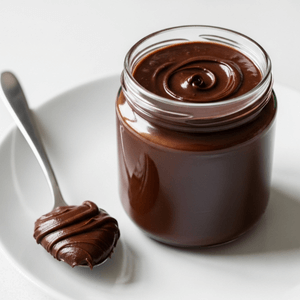 בתמונה צנצנת זכוכית שקופה על צלחת, בתוך הצנצנת ממרח שוקולד ביתי, לצד הצנצנת על הצלחת מונחת כפית כסף ועליה מרח השוקולד