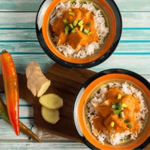 תמונה של שתי צלחות ובהן אורז עם תבשיל עוף בקארי צהבהב וירקות, לצד הצלחות מונחים פלפל חריף ושורש ג׳ינג׳ר פרוס בחלקו